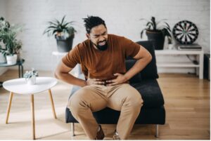 A man sitting on the sofa is suffering from severe IBS disease and is touching his aching abdomen at home